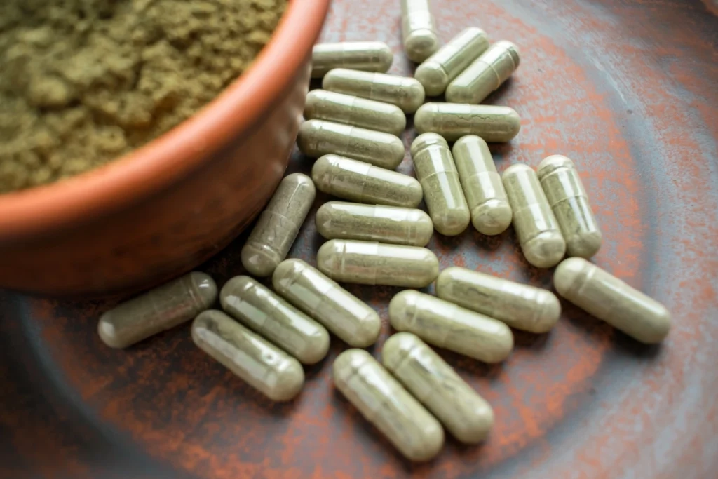 Green kratom powder in a terracotta bowl next to a pile of kratom capsules