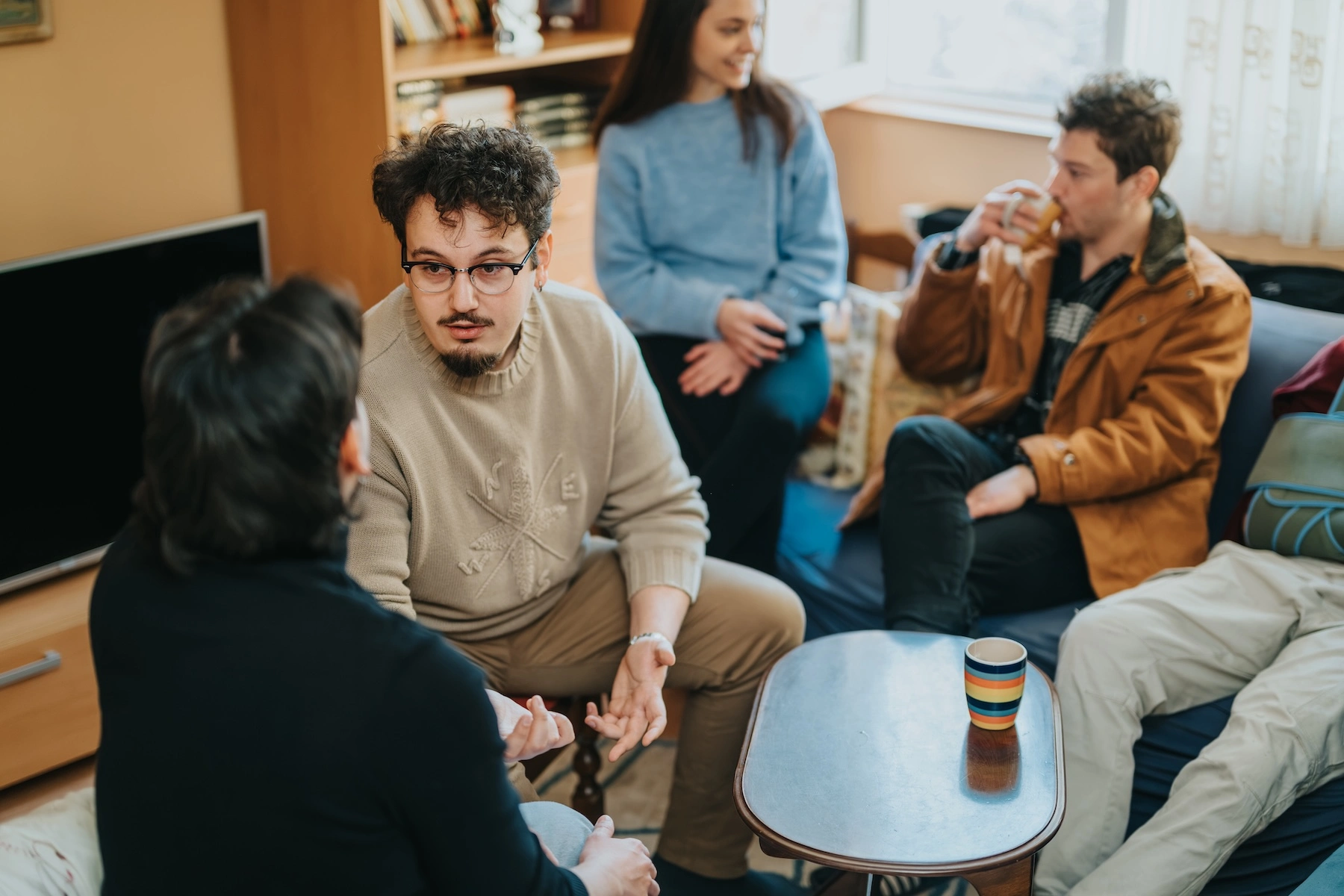 Group of young adults making conversation in a living room as a man in glasses gestures while he talks